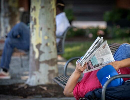 Man laying on a bench reading the newspaper